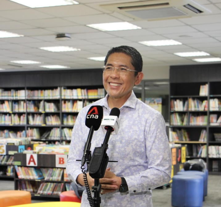 A smiling man in a floral shirt speaks into CNA and Berita microphones inside a library with bookshelves.